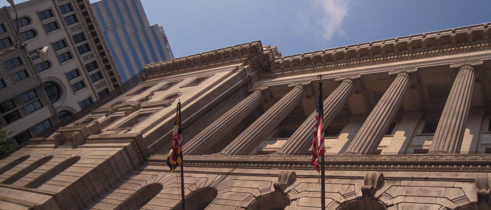 Looking up at the columned stone facade of the Clarence M. Mitchell Jr. Courthouse in Baltimore, with the Maryland and United States flags flying from its front.